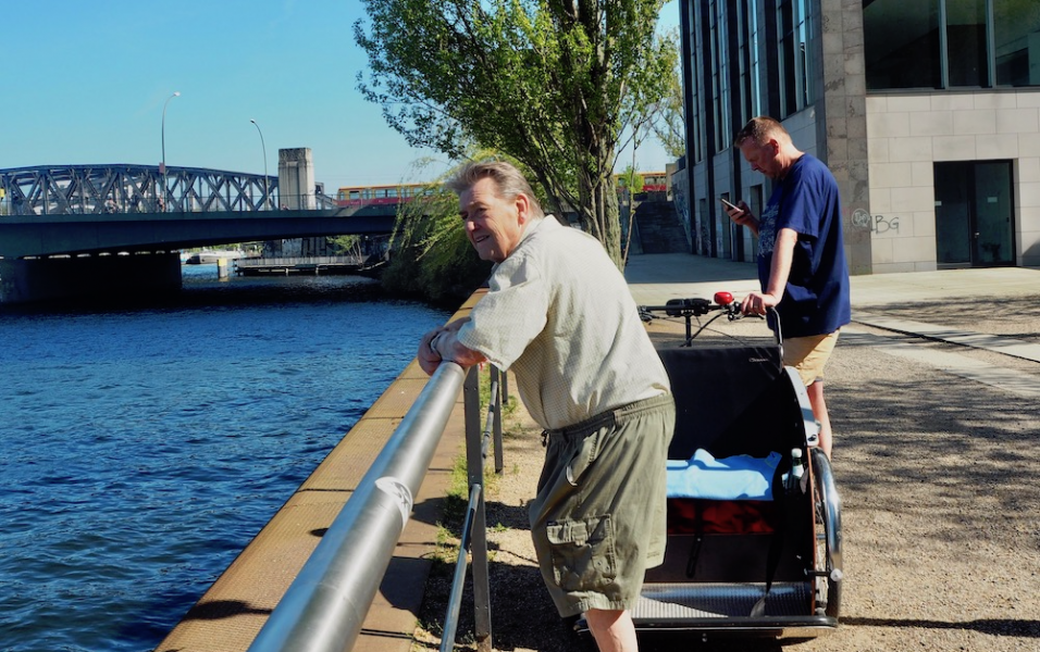 wei Männer stehen am Wasser, zwischen ihnen steht eine parkende Fahrradrikscha